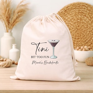 Puede incluir: Una bolsa beige con cordón, con las palabras "Tini Bit Too Fun Mandi's Bachelorette" y una ilustración de una copa de martini. La bolsa es de tela suave y tiene un cierre de cordón.