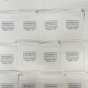 K&ouml;nnte beinhalten: Eine Anordnung von cremefarbenen Stoffbeuteln, jeweils mit "WEDDING RECOVERY KIT", den Namen "MARIA JOSE & JONATHAN" und dem Datum "JANUARY 17, 2026" in Schwarz bedruckt. Die Beutel sind f&uuml;r eine Hochzeit.
