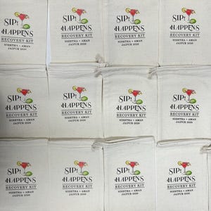 K&ouml;nnte beinhalten: Eine Ansammlung von cremefarbenen Leinens&auml;cken, die jeweils mit den Worten "SIP! HAPPENS" &uuml;ber einer Cocktail-Illustration bedruckt sind. Darunter steht "RECOVERY KIT" und der Name "NISHTHA + AMAN JAIPUR 2026".