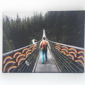 May include: A person walks across a suspension bridge in a forest. The bridge is decorated with rainbow-colored arches.