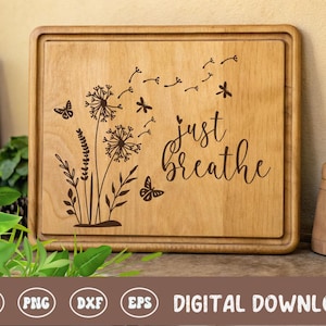 May include: A rectangular wooden cutting board with a carved floral design and the words "just breathe." The design includes dandelions, butterflies, and sprigs of leaves. The board is light brown with a darker brown border.