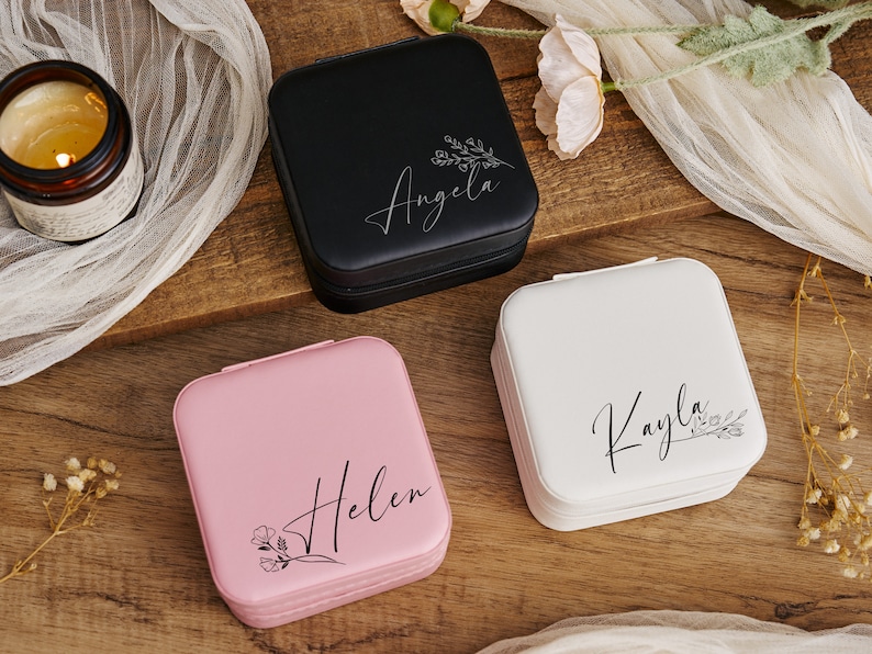 K&ouml;nnte beinhalten: Drei kleine quadratische Schmuckk&auml;stchen mit personalisierten Namen, "Angela", "Kayla" und "Helen", und floralen Designs. Die K&auml;stchen sind schwarz, wei&szlig; und rosa.