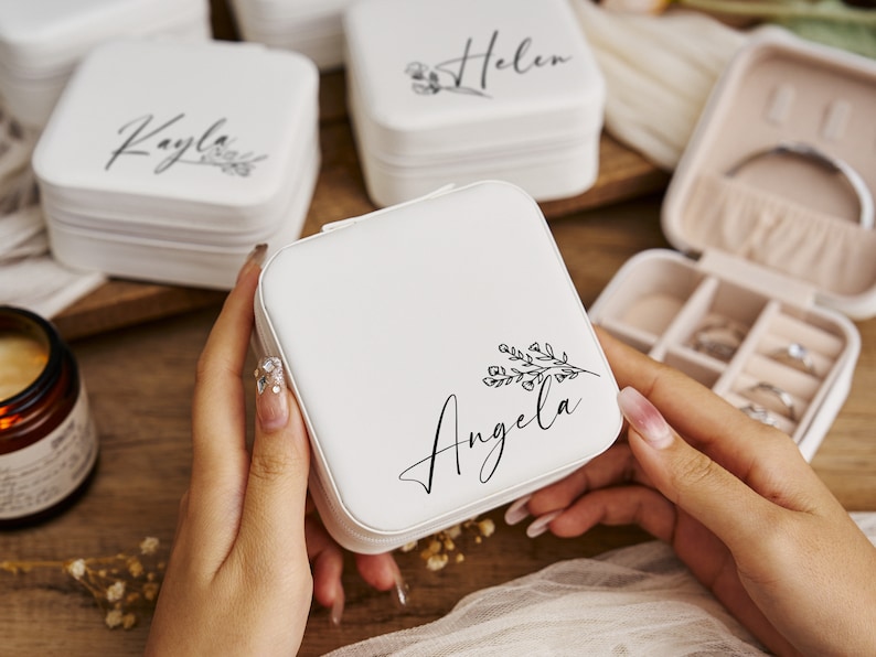 K&ouml;nnte beinhalten: Wei&szlig;e Schmuckschatulle mit floralem Design und dem Namen "Angela" in schwarzer Schrift. Die Schachtel ist ge&ouml;ffnet und hat mehrere F&auml;cher zur Aufbewahrung von Schmuck.