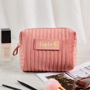 Puede incluir: Una bolsa de maquillaje acolchada de color rosa con una cremallera dorada y una etiqueta dorada que dice "Angela" con un pequeño diseño de flor.