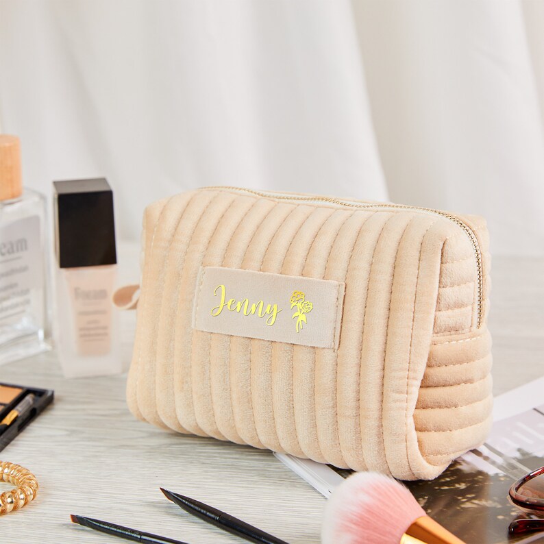 May include: Beige quilted makeup bag with a zipper closure. The bag has a personalised label with the name "Jenny" and a floral design in gold.