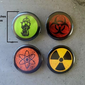 Four circular hazard warning button pins with distressed textures. gas mask silhouette on green. biohazard symbol on red. atom symbol on orange. radiation symbol on yellow. These represent various scientific and safety warning symbols commonly used to indicate dangerous materials or environments. Pins are 1.25" or 32mm round.  Metal Backing. Handmade in the USA. Not safe for children due to the sharp pin on the back. Brought to you by Brain Shudder Studio.