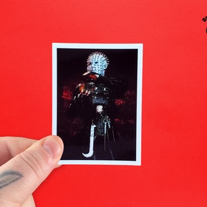 May include: A black and white sticker featuring a character from the horror movie Hellraiser. The character is wearing a spiked helmet and holding a hook. The sticker is on a red background.