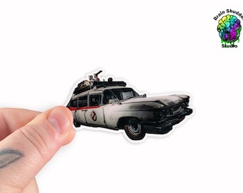May include: A sticker featuring the Ecto-1 from the Ghostbusters film. The white and red vehicle has a detailed design, including equipment on the roof and the Ghostbusters logo. The sticker is held by a hand.