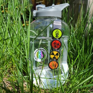 Four circular hazard warning button pins with distressed textures. gas mask silhouette on green. biohazard symbol on red. atom symbol on orange. radiation symbol on yellow. These represent various scientific and safety warning symbols commonly used to indicate dangerous materials or environments. Pins are 1.25" or 32mm round.  Metal Backing. Handmade in the USA. Not safe for children due to the sharp pin on the back. Brought to you by Brain Shudder Studio.