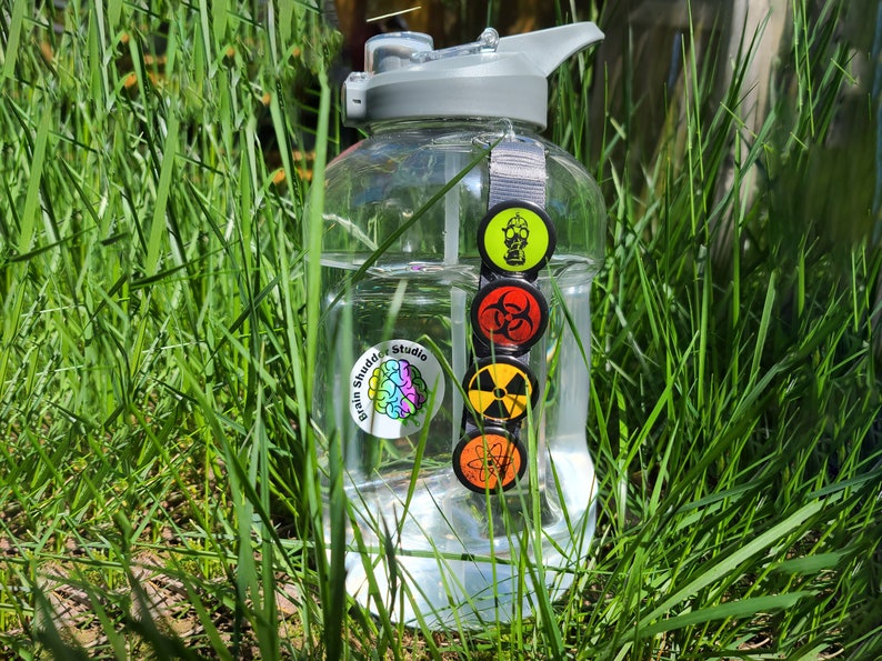 Four circular hazard warning button pins with distressed textures. gas mask silhouette on green. biohazard symbol on red. atom symbol on orange. radiation symbol on yellow. These represent various scientific and safety warning symbols commonly used to indicate dangerous materials or environments. Pins are 1.25" or 32mm round.  Metal Backing. Handmade in the USA. Not safe for children due to the sharp pin on the back. Brought to you by Brain Shudder Studio.