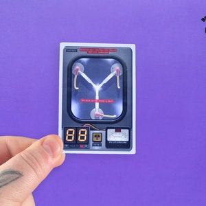 May include: A white sticker with a black outline depicting a futuristic device with a glowing blue light and the text "PLUTONIUM". The device has a digital display showing the number "88".