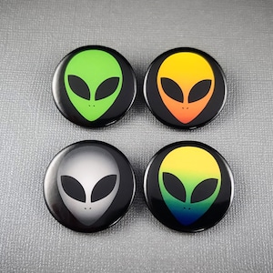 May include: Four round black and white buttons with alien faces. The aliens have green, orange, gray, and rainbow colored faces.