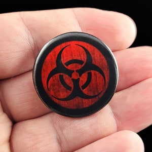 A black biohazard symbol on a distressed red background. Pins are 1.25" or 32mm round.  Metal Backing. Handmade in the USA. Not safe for children due to the sharp pin on the back. Brought to you by Brain Shudder Studio.