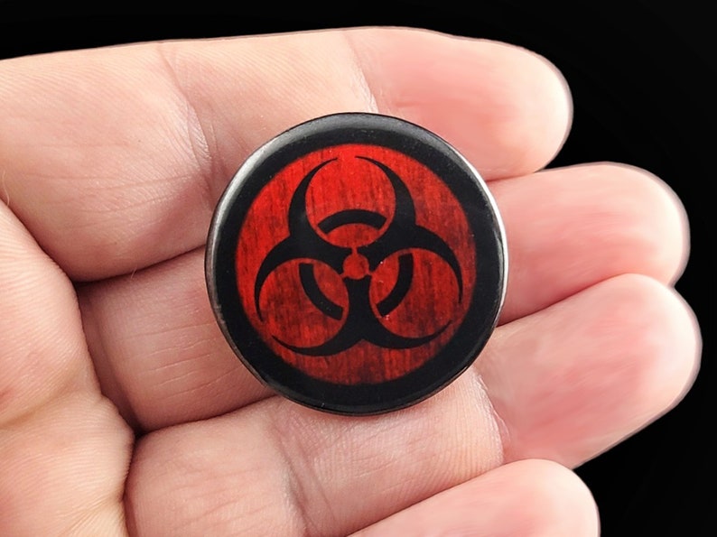 A black biohazard symbol on a distressed red background. Pins are 1.25" or 32mm round.  Metal Backing. Handmade in the USA. Not safe for children due to the sharp pin on the back. Brought to you by Brain Shudder Studio.