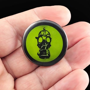 A black gas mask with a green background. Pins are 1.25" or 32mm round.  Metal Backing. Handmade in the USA. Not safe for children due to the sharp pin on the back. Brought to you by Brain Shudder Studio.