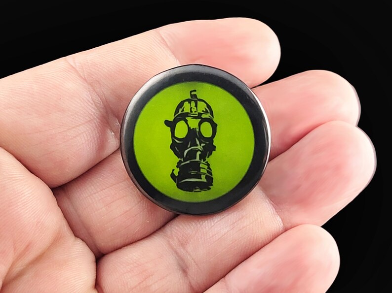 A black gas mask with a green background. Pins are 1.25" or 32mm round.  Metal Backing. Handmade in the USA. Not safe for children due to the sharp pin on the back. Brought to you by Brain Shudder Studio.