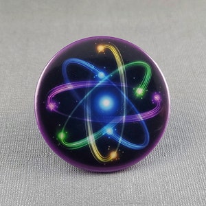 May include: A purple button with a colorful atom design. The atom is made up of a bright blue center with glowing green, yellow, and purple rings around it.