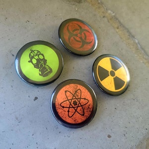 Four circular hazard warning button pins with distressed textures. gas mask silhouette on green. biohazard symbol on red. atom symbol on orange. radiation symbol on yellow. These represent various scientific and safety warning symbols commonly used to indicate dangerous materials or environments. Pins are 1.25" or 32mm round.  Metal Backing. Handmade in the USA. Not safe for children due to the sharp pin on the back. Brought to you by Brain Shudder Studio.