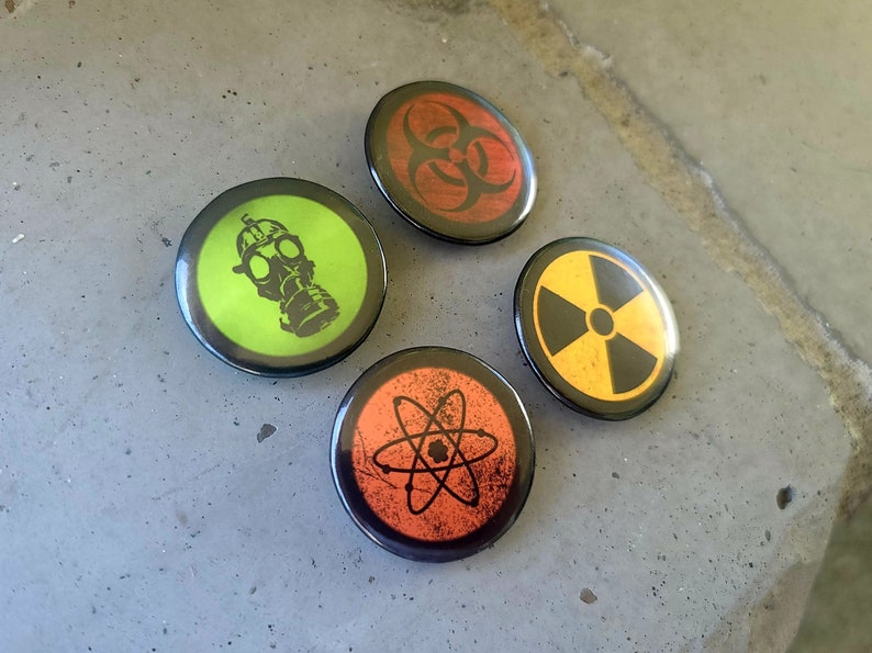 Four circular hazard warning button pins with distressed textures. gas mask silhouette on green. biohazard symbol on red. atom symbol on orange. radiation symbol on yellow. These represent various scientific and safety warning symbols commonly used to indicate dangerous materials or environments. Pins are 1.25" or 32mm round.  Metal Backing. Handmade in the USA. Not safe for children due to the sharp pin on the back. Brought to you by Brain Shudder Studio.
