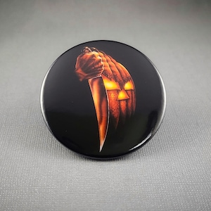May include: A round, black button featuring a carved orange pumpkin with glowing eyes and a knife held in a hand. The design is set against a black background, evoking a Halloween theme.