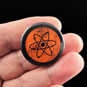 A black and orange button with a black atom symbol on a distressed orange background. Pins are 1.25" or 32mm round.  Metal Backing. Handmade in the USA. Not safe for children due to the sharp pin on the back. Brought to you by Brain Shudder Studio.