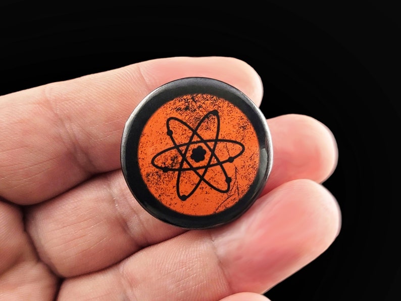 A black and orange button with a black atom symbol on a distressed orange background. Pins are 1.25" or 32mm round.  Metal Backing. Handmade in the USA. Not safe for children due to the sharp pin on the back. Brought to you by Brain Shudder Studio.