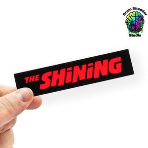 May include: A black and red sticker with the text "THE SHINING" in red letters.