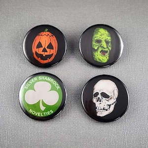 May include: Four black round buttons with Halloween and novelty designs. One button features a jack-o'-lantern, another a green monster face, a third has a shamrock with "Silver Shamrock Novelties", and the last a skull.
