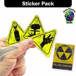 May include: A sticker pack featuring four unique designs. Three stickers are yellow hazard signs with black graphics of a bomb, mushroom cloud, and hand. The fourth sticker is a rectangular sign with a radiation symbol and the text "FALLOUT SHELTER."