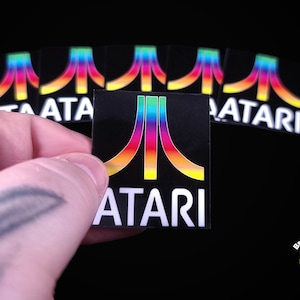 May include: A set of eight rainbow colored Atari video game logo stickers on a black background. The stickers are square and have a white border around the logo. The logo is a stylized "A" with a rainbow colored "I" in the middle.