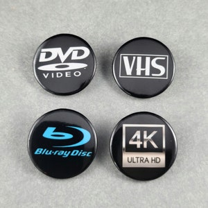 May include: Four black round buttons with different media formats printed in white. The buttons read "DVD VIDEO", "VHS", "Blu-ray Disc", and "4K ULTRA HD".