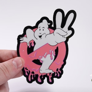 May include: A pink and black sticker featuring a ghost from the Ghostbusters movie making a peace sign. The ghost is dripping pink slime and is in front of the Ghostbusters logo.