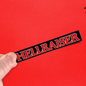 May include: A black and red sticker with the word "HELLRAISER" printed in white letters. The sticker is being held by a hand against a red background.