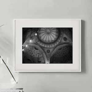 Peut inclure: Photographie en noir et blanc de l'intérieur d'une mosquée, encadrée en blanc. L'image capture les détails complexes du plafond en dôme, avec des motifs ornementaux et des éléments architecturaux. La photo est sur un meuble blanc, avec des livres et un vase.