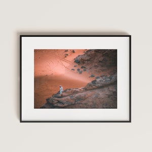 Può includere: Una persona vestita di bianco si trova in piedi su una scogliera rocciosa che domina un vasto deserto di sabbia rossa. Il deserto è illuminato dal caldo bagliore del sole al tramonto, proiettando lunghe ombre sul paesaggio.