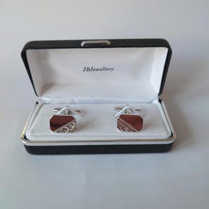 May include: A pair of cufflinks in a black box with the text "JBJewellery". The cufflinks have a rectangular shape with a brown and silver design. The box is open, revealing the cufflinks on a white surface.