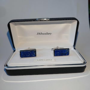 Könnte beinhalten: Rechteckige Manschettenknöpfe mit poliertem Silberfinish und eingelegten Lapislazuli-Steinen, präsentiert in einer schwarzen Schatulle mit dem Text "JBJewellery". Die Schatulle ist mit weißem Stoff ausgekleidet und trägt die Aufschrift "LAPIS LAZULI".