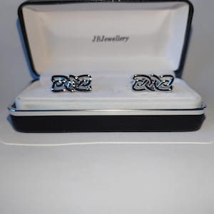 Puede incluir: Gemelos plateados con un intrincado diseño de nudo celta, presentados en una caja de joyería en blanco y negro abierta. Los gemelos descansan sobre un forro de satén blanco. El nombre de la marca "JBJewellery" está impreso en el interior.