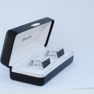 Könnte beinhalten: Ein Paar schwarze und silberne Manschettenknöpfe in einer schwarzen Lederbox mit dem Text "JB Jewellery" auf der Innenseite des Deckels.