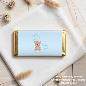 May include: A rectangular chocolate bar wrapped in a light blue paper sleeve featuring a brown teddy bear holding three balloons. The text reads We can Bearly Wait and BABY WARREN IS ON THE WAY. The ends of the bar are covered in shiny gold foil.