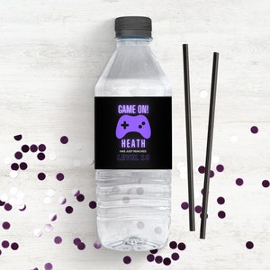 Puede incluir: Una etiqueta de botella de agua negra y morada con un gráfico de mando de videojuego y el texto "GAME ON! HEATH HAS JUST REACHED LEVEL 30".
