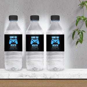 May include: Three clear plastic water bottles with black labels featuring a blue video game controller graphic and the text "GAME ON!" and "HEATH HAS JUST REACHED LEVEL 20". The bottles have black caps.