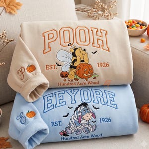 May include: A beige sweatshirt featuring Winnie the Pooh dressed as a bee with a pumpkin and a light blue sweatshirt featuring Eeyore, both with collegiate-style text reading POOH or EEYORE, EST. 1926, and Hundred Acre Wood, plus pumpkin sleeve embroidery.