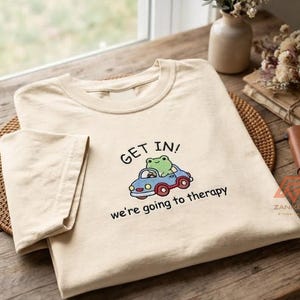 Può includere: Una t-shirt a girocollo color beige con la grafica di una rana verde alla guida di una piccola auto blu con ruote rosse. Il testo sopra l'auto recita "GET IN!" e il testo sotto l'auto recita "we're going to therapy" in carattere nero.