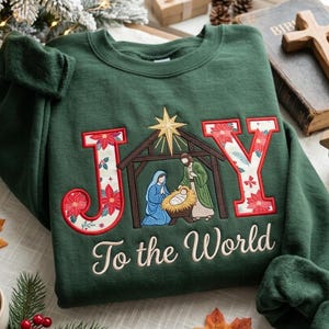May include: A dark green crewneck sweatshirt featuring the word JOY embroidered in large letters. The J and Y are filled with a red poinsettia floral pattern, while the O is replaced by a nativity scene graphic. The text To the World is below.