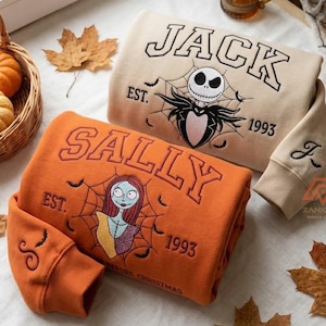 May include: A beige sweatshirt featuring a Jack Skellington graphic with the text JACK EST. 1993 and an orange sweatshirt featuring a Sally graphic with the text SALLY EST. 1993. Both garments have embroidered details on the sleeves and cuffs.