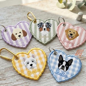Puede incluir: Cinco llaveros de tela en forma de corazón con cierres de metal dorado o plateado. Cada uno presenta un retrato de perro bordado y un nombre: Coco sobre rayas lavanda, Jasper sobre rayas verdes, Sunny sobre rayas rosas, Bodie sobre cuadros vichy amarillos y Stella sobre cuadros vichy azules.