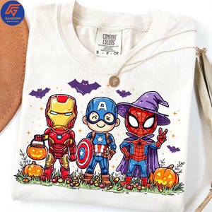 May include: Cream-coloured t-shirt featuring cartoon superheroes Iron Man, Captain America, and Spider-Man in Halloween costumes. The shirt includes pumpkins, bats, and the Comfort Colors brand label. A gold necklace is draped across the shirt.