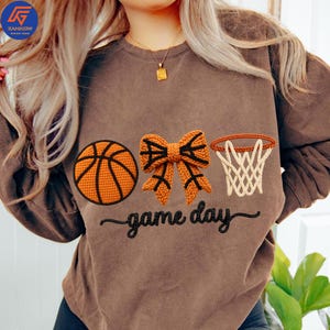 May include: Brown sweatshirt with embroidered basketball, bow, and hoop designs. The text "game day" is embroidered below the graphics. The sweatshirt has long sleeves and a crew neck.