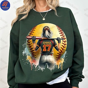 K&ouml;nnte beinhalten: Gr&uuml;nes Sweatshirt mit einem Aquarell-Design einer Softballspielerin, die einen Schl&auml;ger h&auml;lt. Die Spielerin tr&auml;gt ein schwarz-rotes Trikot mit dem Namen "EMMA" und der Nummer "17" auf dem R&uuml;cken.